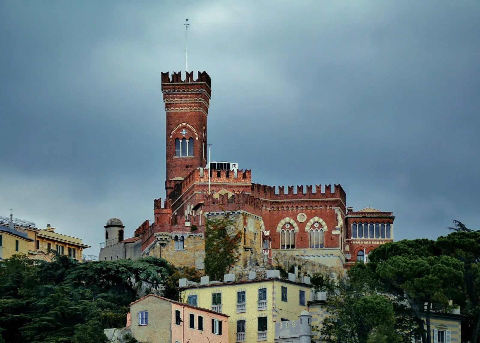 Il Castello d'Albertis in mattoni rossi con la torre merlata, sopra le case colorate della collina di Montegalletto sotto un cielo nuvoloso