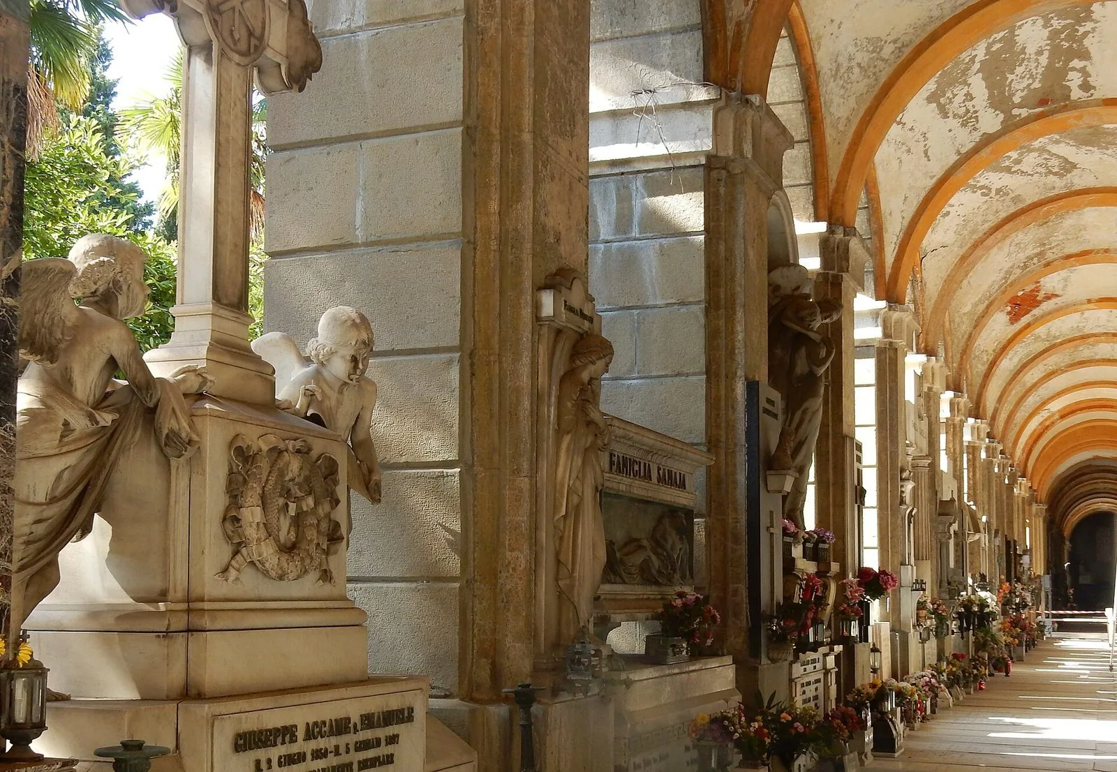 Un porticato del Cimitero monumentale di Staglieno con volte ad arco, angeli e statue in marmo lungo le tombe e fiori ai lati